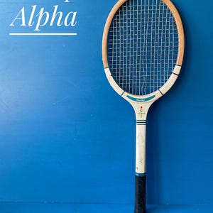 A vintage Dunlop Alpha tennis racket with a wooden frame. The handle has a dark black grip. The text "Dunlop Alpha" is displayed in white script on a blue background.