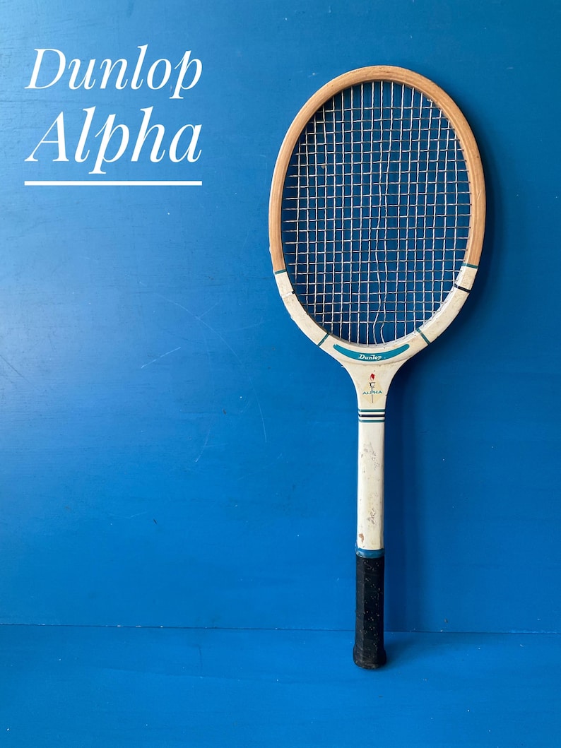 A vintage Dunlop Alpha tennis racket with a wooden frame. The handle has a dark black grip. The text "Dunlop Alpha" is displayed in white script on a blue background.