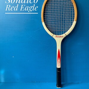 May include: A vintage wooden tennis racket labeled Sondico Red Eagle. The racket features a natural wood frame, white throat, and a black handle grip. The strings are light-colored, and the shaft displays a red graphic design with the text Red Eagle.