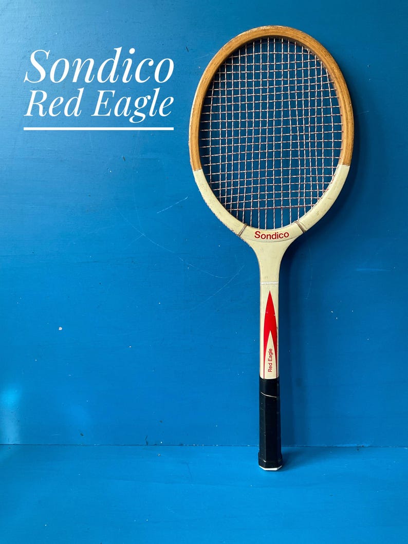 May include: A vintage wooden tennis racket labeled Sondico Red Eagle. The racket features a natural wood frame, white throat, and a black handle grip. The strings are light-colored, and the shaft displays a red graphic design with the text Red Eagle.