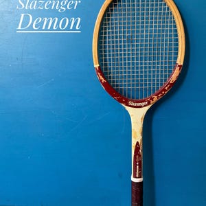 A vintage wooden Slazenger Demon tennis racket. The racket is predominantly beige and burgundy. The racket is photographed against a blue background