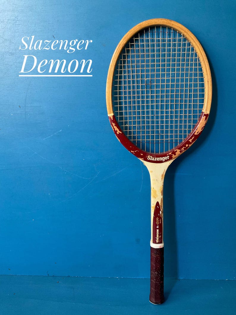 A vintage wooden Slazenger Demon tennis racket. The racket is predominantly beige and burgundy. The racket is photographed against a blue background