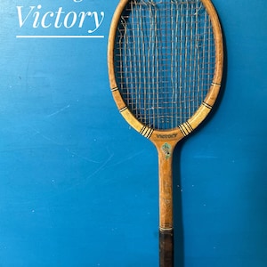 A vintage wooden tennis racket with the text Slazenger Victory printed on the frame. The racket features a natural wood finish, a dark brown grip, and worn, frayed strings. A small decorative emblem is visible near the throat.