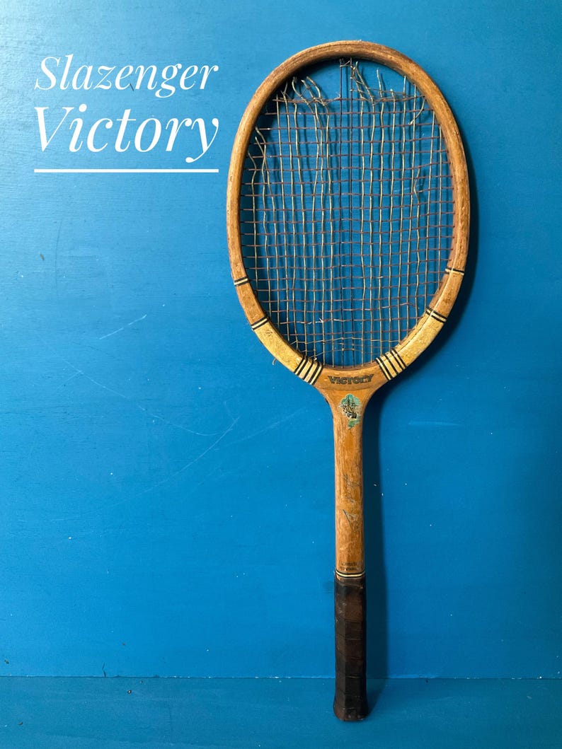 A vintage wooden tennis racket with the text Slazenger Victory printed on the frame. The racket features a natural wood finish, a dark brown grip, and worn, frayed strings. A small decorative emblem is visible near the throat.