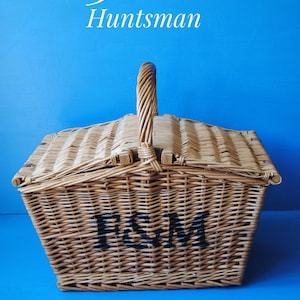 Large Fortnum and Mason willow Huntsman style picnic basket. The basket is sitting on a blue surface, with the Fortnum and Mason logo visible at the top. The basket has an integrated willow handle to the front, plus a hinged lid that opens from the middle