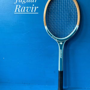 A vintage wooden tennis racket by Jaguar. The racket has a blue and white shaft, with natural wood coloured head. It has a dark handgrip, and is photographed against a blue background