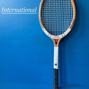 A vintage wooden tennis racket featuring a light brown wood frame with white accents, white nylon strings, and a black grip. The word International is printed in white on the left and in black on the throat of the racket with a red star.
