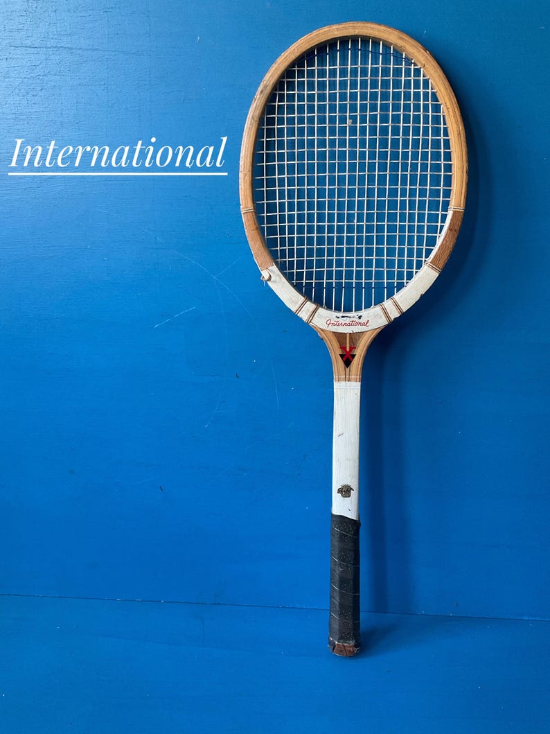 A vintage wooden tennis racket featuring a light brown wood frame with white accents, white nylon strings, and a black grip. The word International is printed in white on the left and in black on the throat of the racket with a red star.