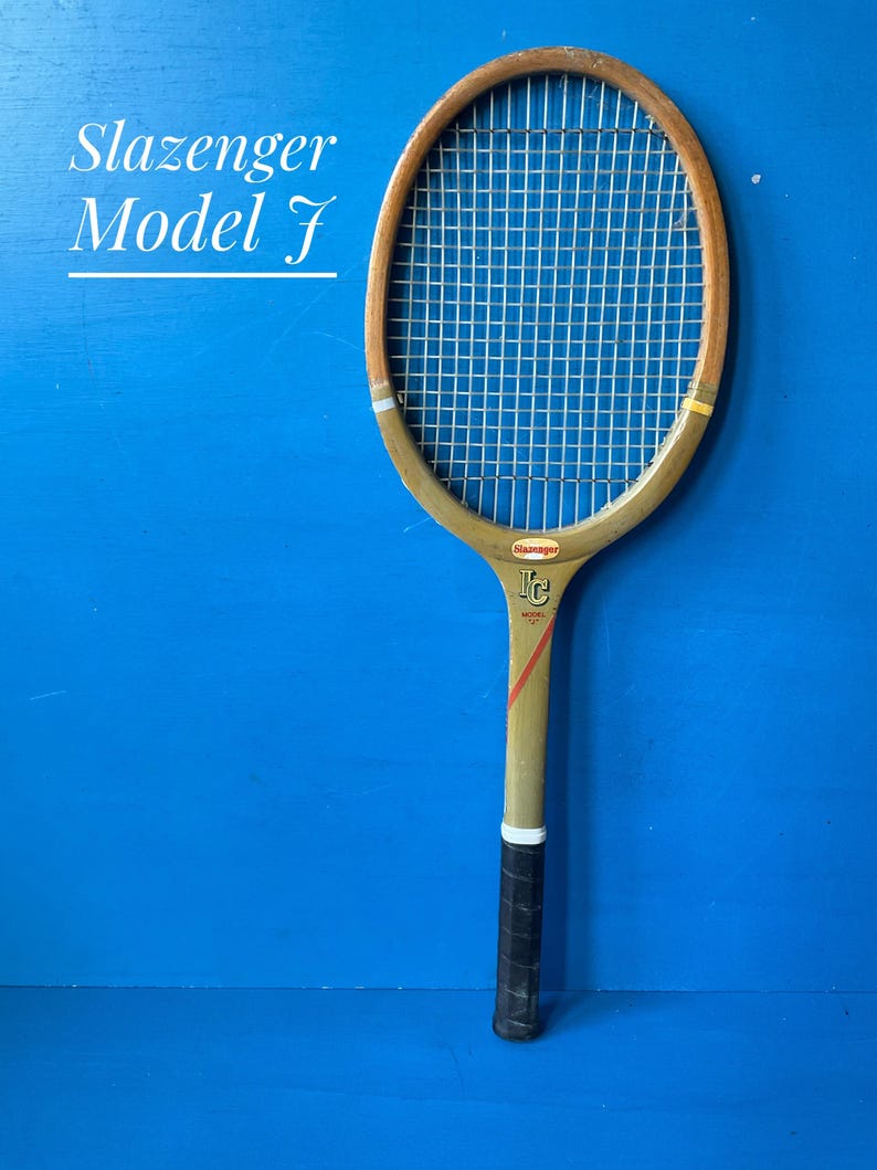 A vintage Slazenger Model J wooden tennis racket. The racket is a faded olive green, with the slazenger logo in red. The top half of the racket head is in natural wood. The racket has a dark leather hand grip