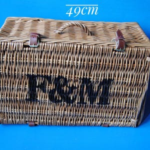 49cm Fortnum and Mason willow picnic basket. The basket is sitting upright, with the Fortnum and Mason logo facing front. The basket has an integrated willow handle, plus a hinged lid that has two leather straps to hold the lid securely in-place