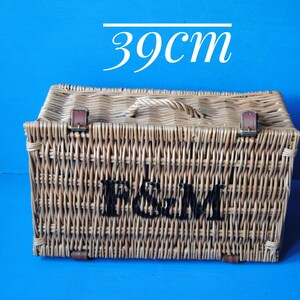 39cm Fortnum and Mason willow picnic basket. The basket is sitting upright, with the Fortnum and Mason logo facing front. The basket has an integrated willow handle, plus a hinged lid that has two leather straps to hold the lid securely in-place