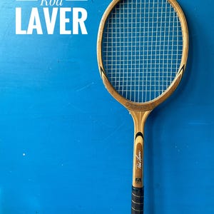 A vintage wooden tennis racket with a natural wood frame, white painted accents, and a brown leather grip. The racket features the text Rod Laver printed on the frame and the head, with a standard nylon string pattern.