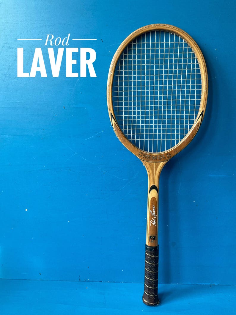 A vintage wooden tennis racket with a natural wood frame, white painted accents, and a brown leather grip. The racket features the text Rod Laver printed on the frame and the head, with a standard nylon string pattern.