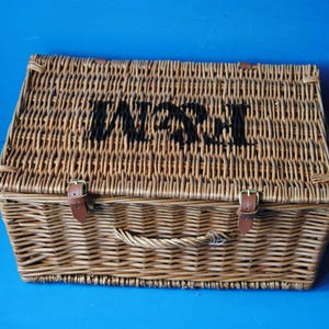 51cm Fortnum and Mason willow picnic basket. The basket is sitting on a blue surface, with the Fortnum and Mason logo visible at the top. The basket has an integrated willow handle to the front, plus a hinged lid that has two leather straps to hold the lid securely in-place