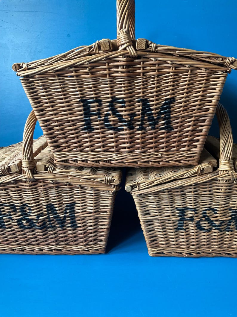 Three Fortnum and Mason Huntsman style wicker baskets with bi- fold lids and wicker handle. The baskets are stacked, with the F&M logo facing forward