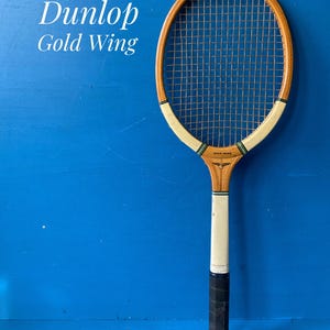 May include: A vintage Dunlop Gold Wing wooden tennis racket featuring a natural wood frame, white throat accents with green and black stripes, a black grip, and a standard string pattern. The text Dunlop Gold Wing is visible on the frame.