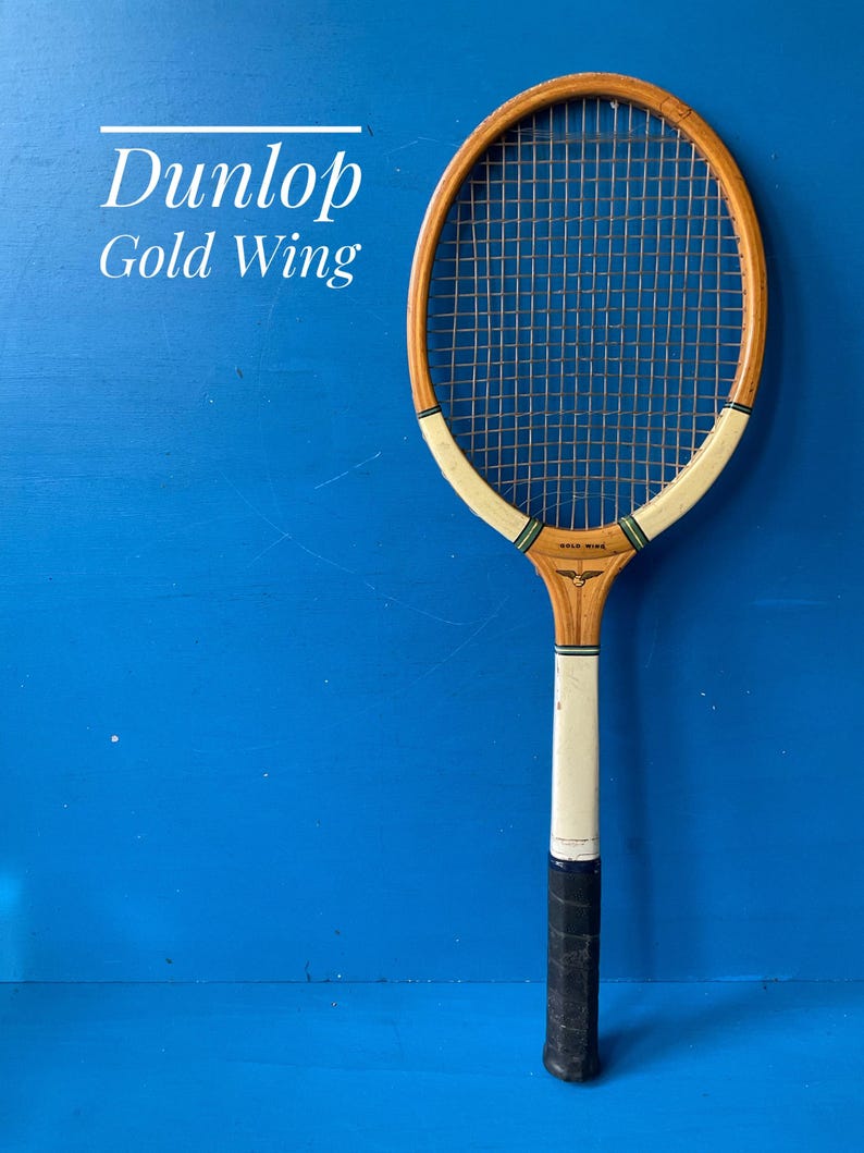 May include: A vintage Dunlop Gold Wing wooden tennis racket featuring a natural wood frame, white throat accents with green and black stripes, a black grip, and a standard string pattern. The text Dunlop Gold Wing is visible on the frame.