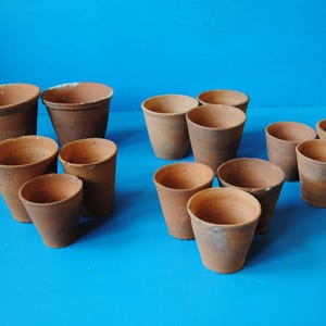 May include: A collection of thirteen small, unglazed terracotta clay cups with a tapered, conical shape and a natural reddish-brown finish. Each cup features a simple, open rim and a smooth, matte texture, arranged in a loose grouping.