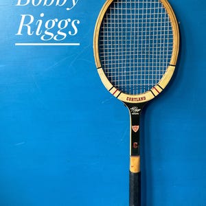 A vintage wooden tennis racket with white strings, a black handle, and the name "Cortland" on the frame. The racket has red and black accents. The name "Bobby Riggs" is written on the blue background.