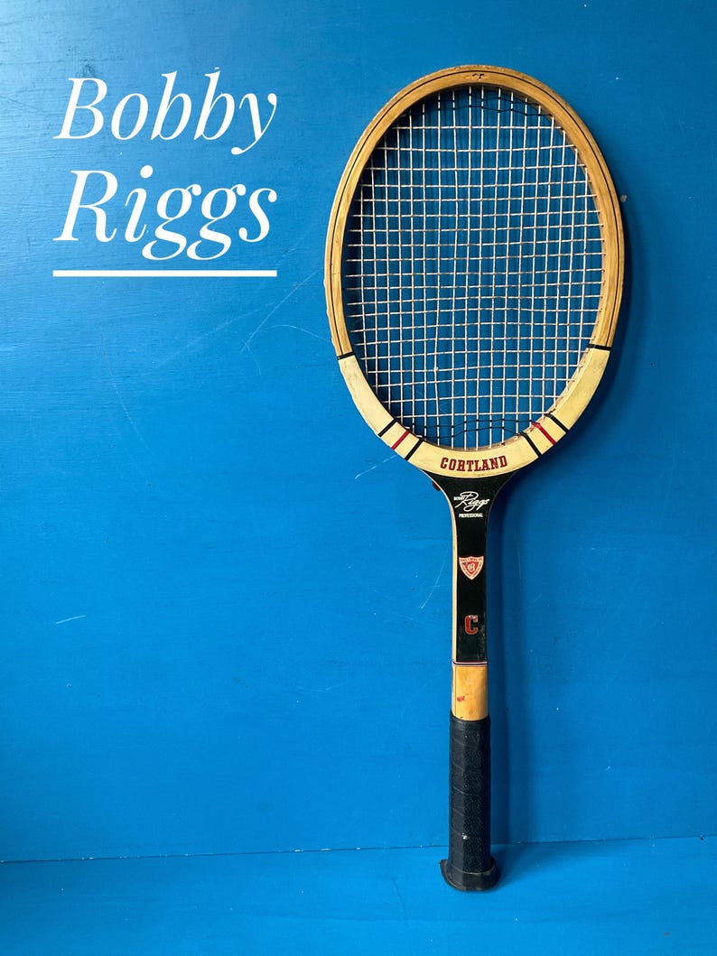 A vintage wooden tennis racket with white strings, a black handle, and the name "Cortland" on the frame. The racket has red and black accents. The name "Bobby Riggs" is written on the blue background.