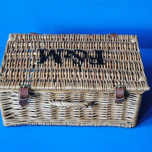 39cm Fortnum and Mason willow picnic basket. The basket is sitting on a blue surface, with the Fortnum and Mason logo visible at the top. The basket has an integrated willow handle to the front, plus a hinged lid that has two leather straps to hold the lid securely in-place