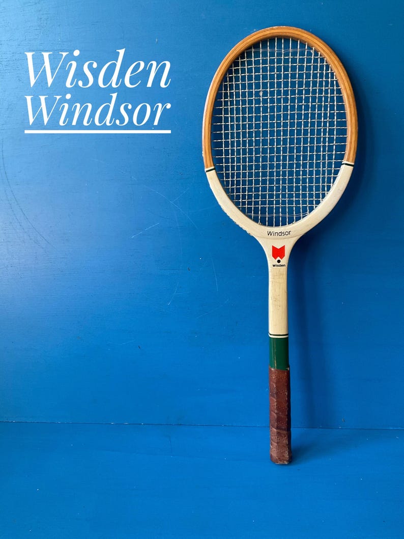 A vintage wooden tennis racket featuring a light wood frame, white throat, and a brown leather grip. The racket includes a red logo and the text "Wisden" and "Windsor" on the frame. The text "Wisden Windsor" is also visible in white.