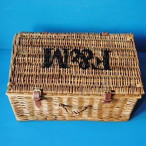 49cm Fortnum and Mason willow picnic basket. The basket is sitting on a blue surface, with the Fortnum and Mason logo visible at the top. The basket has an integrated willow handle to the front, plus a hinged lid that has two leather straps to hold the lid securely in-place