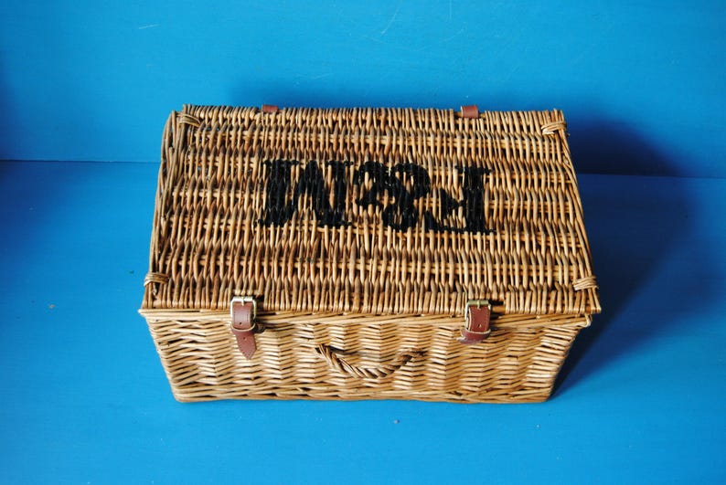 49cm Fortnum and Mason willow picnic basket. The basket is sitting on a blue surface, with the Fortnum and Mason logo visible at the top. The basket has an integrated willow handle to the front, plus a hinged lid that has two leather straps to hold the lid securely in-place