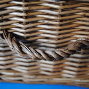 Close-up of a woven wicker Fortnum and Mason hamper basket with a twisted handle. The basket is made of light brown wicker and has a textured surface. The handle is made of tightly wound strands of the same material.