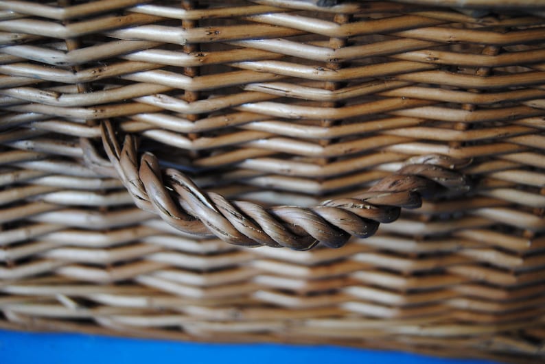 Close-up of a woven wicker Fortnum and Mason hamper basket with a twisted handle. The basket is made of light brown wicker and has a textured surface. The handle is made of tightly wound strands of the same material.