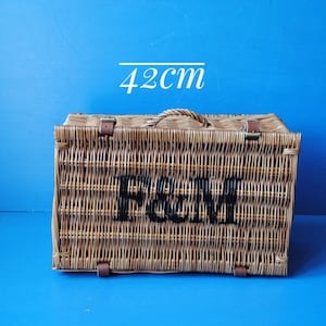 42cm Fortnum and Mason willow picnic basket. The basket is sitting on a blue surface, with the Fortnum and Mason logo visible at the top. The basket has an integrated willow handle to the front, plus a hinged lid that has two leather straps to hold the lid securely in-place