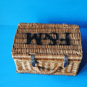 32cm Fortnum and Mason willow picnic basket. The basket is sitting on a blue surface, with the Fortnum and Mason logo visible at the top. The basket has an integrated willow handle to the front, plus a hinged lid that has two leather straps to hold the lid securely in-place