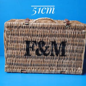 51cm Fortnum and Mason willow picnic basket. The basket is sitting upright, with the Fortnum and Mason logo facing front. The basket has an integrated willow handle, plus a hinged lid that has two leather straps to hold the lid securely in-place