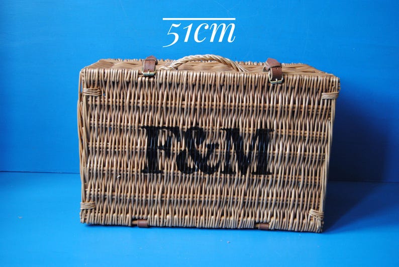 51cm Fortnum and Mason willow picnic basket. The basket is sitting upright, with the Fortnum and Mason logo facing front. The basket has an integrated willow handle, plus a hinged lid that has two leather straps to hold the lid securely in-place