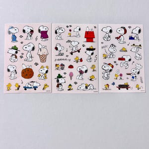 May include: Three sheets of Snoopy stickers showing Snoopy in different poses and activities. Snoopy is shown with an ice cream cone, on a skateboard, and wearing a chef's hat. The stickers are on a light pink background with paw prints.