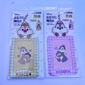 May include: Two photo card holders featuring Chip and Dale from Disney. The card holders are made of plastic and have a checkered pattern. The left card holder is yellow with a brown checkered pattern and the right card holder is pink with a purple checkered pattern. Both card holders have a photo frame in the centre and a small picture of Chip and Dale holding an acorn. The text "DALE" is printed on the left card holder and the text "CHIP" is printed on the right card holder.