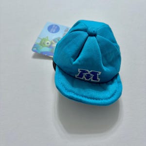 Puede incluir: Una gorra de béisbol de color turquesa brillante con una letra M bordada en color morado oscuro en el panel frontal. La gorra tiene una corona estructurada con un botón en la parte superior y una visera curva. Incluye una etiqueta rectangular.