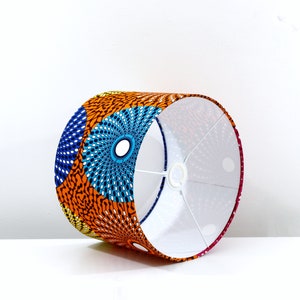 May include: A cylindrical lampshade with a white interior and an exterior covered in colorful African print fabric. The fabric features a geometric pattern of orange, blue, and white circles.