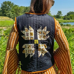 May include: A black quilted vest with a large patchwork design on the back. The design features a yellow, white, and brown geometric pattern. The vest is worn over a striped orange and white dress.