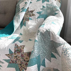May include: A handmade quilt with a starburst pattern in white, teal, and floral fabrics. The quilt features intricate quilting details and a teal border. The starbursts are made of white fabric with teal accents and floral centers.