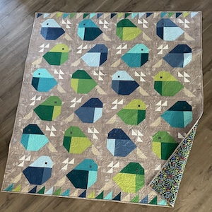 Peut inclure: Une courtepointe avec un motif géométrique d'oiseaux bleus, verts et blancs sur un fond beige. La courtepointe est pliée en deux et le verso est visible, qui est un motif floral dans les tons bleus et verts.