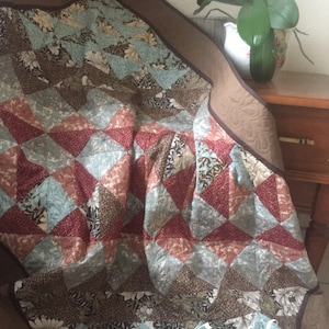 Peut inclure: Une courtepointe de couleur marron, bleue et rouge avec un motif en losange. La courtepointe est pliée et a une bordure marron. Le texte "Sew Busy" est visible dans le coin inférieur droit de l'image.