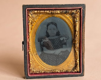 May include: Antique framed portrait of a young person. The oval portrait is set within an ornate gold-colored frame, surrounded by a red border, and encased in a black rectangular case. The portrait depicts a person seated in a chair, wearing a patterned dress.
