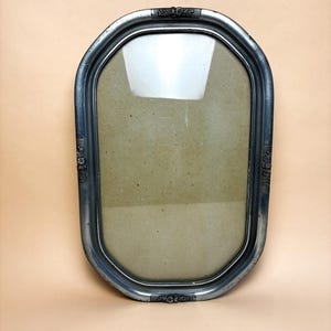 May include: Oval-shaped silver picture frame with a glass front. The frame has decorative floral accents at the top and bottom, and on the sides. The frame is empty, with a tan backing visible through the glass.