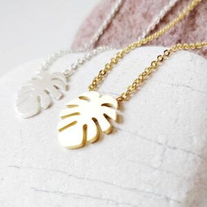 Puede incluir: Dos collares de oro y plata con un colgante de hoja de monstera. Los collares est&aacute;n sobre un fondo de piedra blanca.