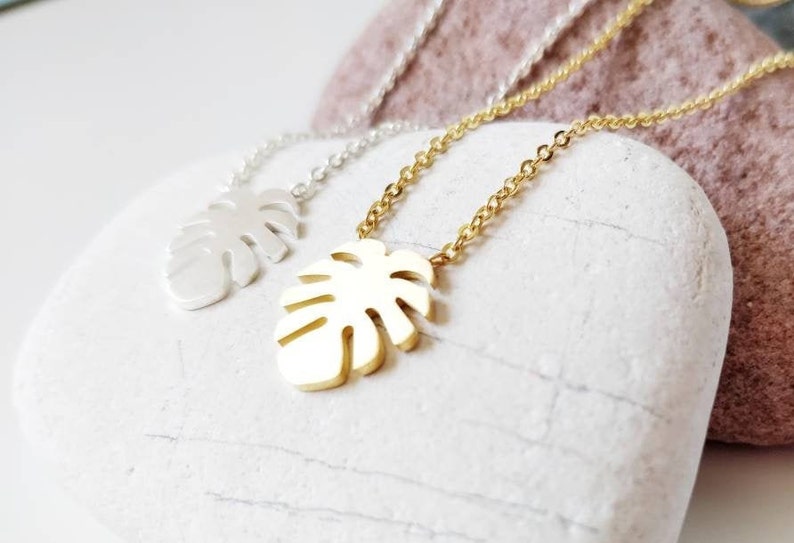 Puede incluir: Dos collares de oro y plata con un colgante de hoja de monstera. Los collares est&aacute;n sobre un fondo de piedra blanca.
