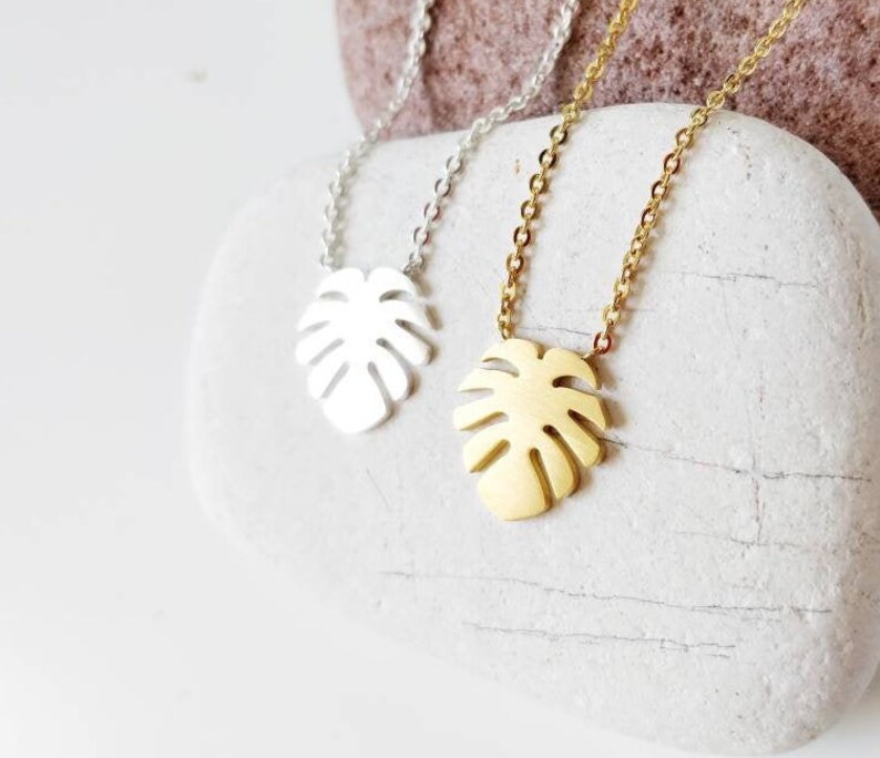 Puede incluir: Dos delicados collares con un colgante de hoja tropical. Un collar es plateado y el otro es dorado.