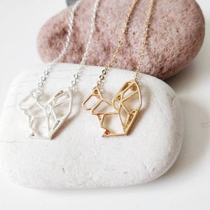May include: Two delicate necklaces with geometric heart pendants. One necklace has a silver pendant, and the other has a gold pendant. Both necklaces are displayed on a white stone.