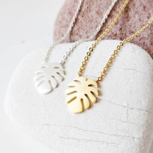 Puede incluir: Dos delicados collares con un colgante de hoja tropical. Un collar es plateado y el otro es dorado. Los collares se muestran sobre una piedra blanca.
