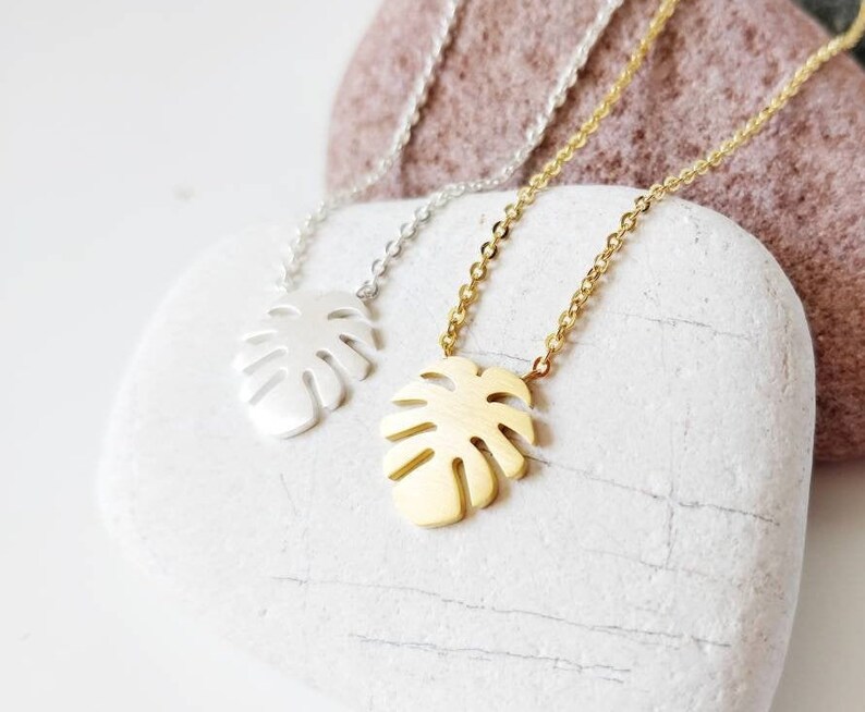 Puede incluir: Dos delicados collares con un colgante de hoja tropical. Un collar es plateado y el otro es dorado. Los collares se muestran sobre una piedra blanca.